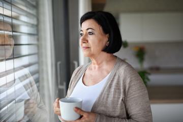Sad senior woman standing and looking out through window indoors at home.