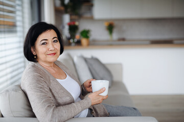 Happy senior woman with coffee sitting on sofam and looking at camera indoors at home.