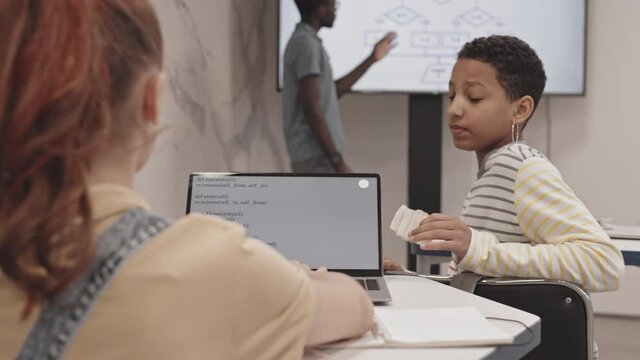 Rear view of teenage Black girl sitting at desk in classroom at lesson, turning around to female Caucasian friend, helping her with assignment, pointing at computer code on laptop screen