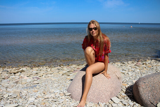 A beautiful, long-legged blonde girl in sunglasses and a red shirt with her hair down is sitting on a large rock, one leg bent, on the beach.
