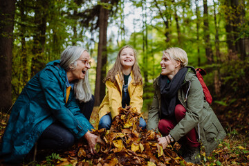 Happy little girl with mother and grandmother having fun with leaves during autumn walk in forest