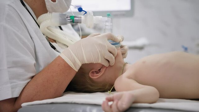 child health, male kid nurse wearing gloves, holds an oxygen mask on his face to inject little patient into anesthesia before heart surgery in a hospital ward