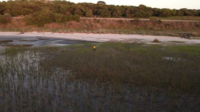 Aerial view of man with taking photos with mobile phone between wet grass at lagoon shore during sunset time - Black Lagoon,Uruguay