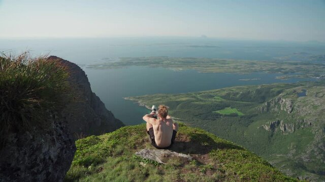 Shirtless Man Hiker Sitting On A Rocky Ledge Taking Travel Photos Using A Long Lens Camera At Mount Donnamannen, Nordland, Norway. - Wide Shot