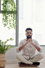 Calm relaxed man sitting on small pillow on the floor and connecting fingers when meditating