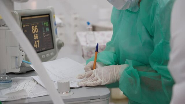 modern medicine, doctor in protective suit, mask and gloves writes the diagnosis in the patient's outpatient card background of monitor connected to person during an operation