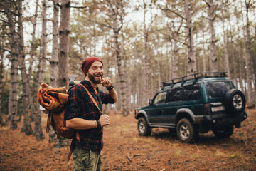 Millennial man hipster with beard wearing backpack, holding axe and firewood in forest with car on...