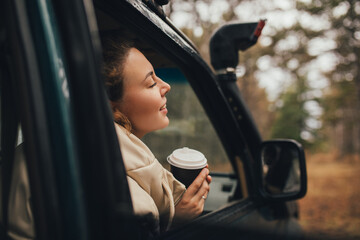 Millennial woman travelling off-road by car in a forest, drinking coffee, enjoying wind. Wanderlust concept.