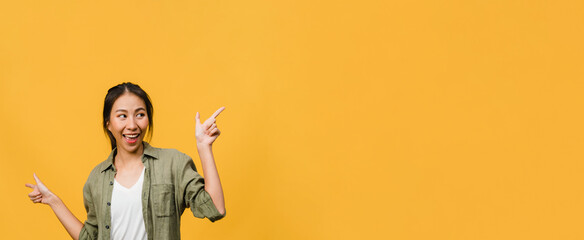 Portrait of young Asian lady smiling with cheerful expression, shows something amazing at blank space in casual clothing and standing isolated over yellow background. Panoramic banner with copy space. - obrazy, fototapety, plakaty
