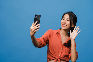 Smiling adorable Asian female making selfie photo on smart phone with positive expression in casual clothing and stand isolated on blue background. Happy adorable glad woman rejoices success.