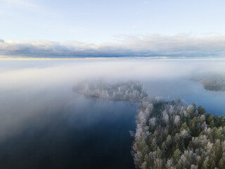 Aerial view over frozen lake in a misty forest, Finland. Winter season. View from above water surface, frozen trees and low fog clouds landscape...