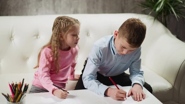 Little blonde girl looks at friend student writing on paper sheet to copy homework studying together at low table in living room closeup slow motion