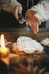 Woman in apron decorating christmas gingerbread cookies with sugar powder on rustic wooden table with candle and ornaments. Christmas holiday preparation and traditions. Atmospheric moody image
