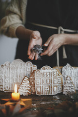 Woman in apron sprinkling sugar powder on christmas gingerbread houses on wooden table in rustic scandinavian kitchen. Atmospheric moody image. Christmas holiday preparation and traditions