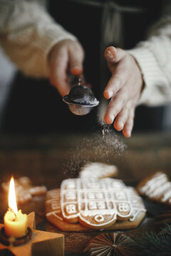 Woman in apron decorating christmas gingerbread cookies with sugar powder on rustic wooden table with candle and ornaments. Christmas holiday preparation and traditions. Moody image