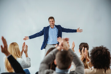 Smiling motivational speaker standing in front of his audience who is clapping. - obrazy, fototapety, plakaty