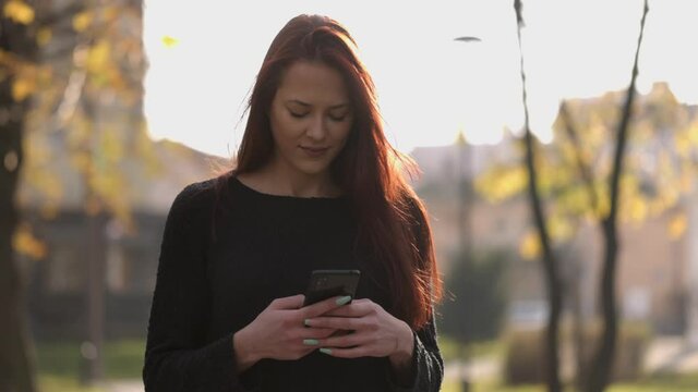 Portrait of happy hipster woman typing by mobile phone outdoors. Closeup cheerful girl stanging with smartphone in city park. Smiling lady holding cellphone in hands outside.
