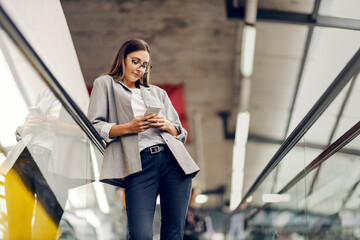 A young businesswoman leaning on the railing while typing an important message on the phone. Busy woman descending escalator and typing message. A businesswoman using the phone
