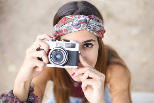 Portrait of a caucasian young brunette with colored ribbon in her hair, posing and making pictures using a retro camera outdoors in park.