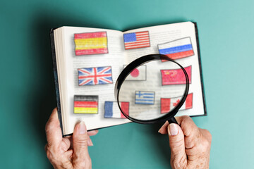 A senior woman is holding a magnifying glass and a book with flags of different countries. Close...