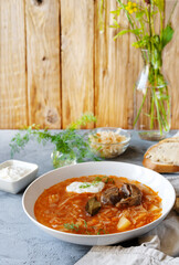 Sour cabbage soup with ingredients in a white plate, wooden background and grey table