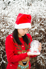 An asian korean girl in red jumper and sants hat holding a snow globe in the snow