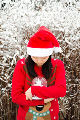 An asian korean girl in red jumper and sants hat holding a snow globe in the snow