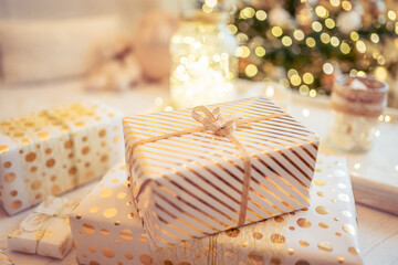 A lot of packing handmade gift boxes lying on the table near Christmas tree in the midst of golden lights, glowing garland, candle. Soft focus