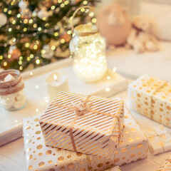 A lot of packing handmade gift boxes lying on the table near Christmas tree in the midst of golden lights, glowing garland, candle. Soft focus. square