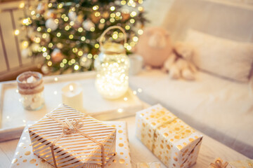 Obraz na płótnie Canvas A lot of packing handmade gift boxes lying on the table near Christmas tree in the midst of golden lights, glowing garland, candle. Soft focus