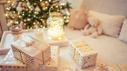 Fototapeta na wymiar A lot of packing handmade gift boxes lying on the table near Christmas tree in the midst of golden lights, glowing garland, candle. Soft focus. 16:9