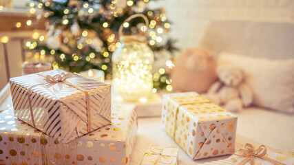 Fototapeta na wymiar A lot of packing handmade gift boxes lying on the table near Christmas tree in the midst of golden lights, glowing garland, candle. Soft focus. 16:9