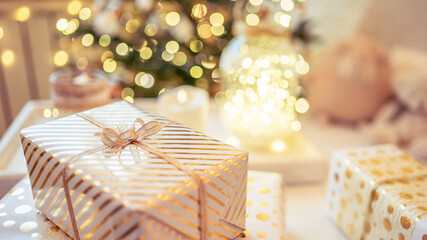 A lot of packing handmade gift boxes lying on the table near Christmas tree in the midst of golden lights, glowing garland, candle. Soft focus. 16:9