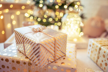 Fototapeta na wymiar A lot of packing handmade gift boxes lying on the table near Christmas tree in the midst of golden lights, glowing garland, candle. Soft focus