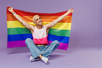 Full size young latin gay man with make up wearing bright pink top sitting hold rainbow striped flag look aside isolated on plain pastel purple background studio People lifestyle fashion lgbtq concept
