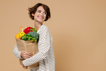 Young smiling cheerful vegetarian woman 20s in casual clothes hold paper bag with vegetables...