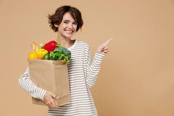 Young smiling vegetarian woman in casual clothes hold paper bag with vegetables after shopping in greengrocery point finger aside on workspace area isolated on plain pastel beige background studio.