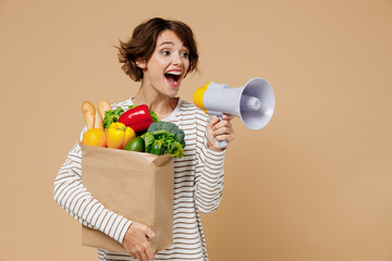 Young smiling vegetarian woman in casual clothes hold paper bag with vegetables hold scream in...