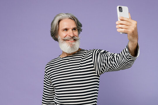Elderly gray-haired mustache bearded man 50s wearing striped turtleneck doing selfie shot on mobile cell phone post photo on social network isolated on plain pastel light purple background studio