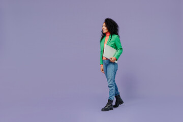 Full size body length charismatic fun young black curly woman 20s wears casual clothes hold laptop pc computer under hand go move walk isolated on plain pastel light violet background studio portrait.