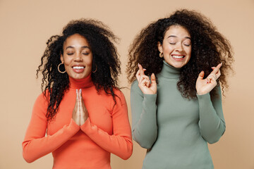 Two young curly black women friends 20s wearing casual shirts clothes holding hands folded in prayer begging making wish keep fingers crossed isolated on plain pastel beige background studio portrait.
