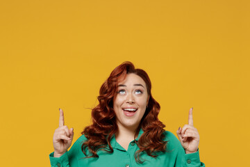 Bright happy vivid young ginger chubby overweight woman 20s wears green shirt pointing index finger up above on workspace area copy space mock up isolated on plain yellow background studio portrait.