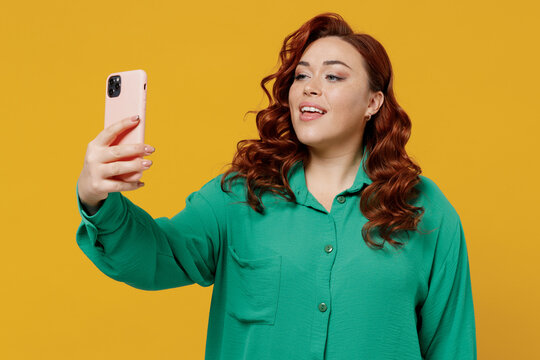 Bright happy vivid young ginger chubby overweight woman 20s wears green shirt doing selfie shot on mobile cell phone post photo on social network isolated on plain yellow background studio portrait.