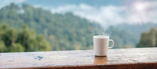 White mug of hot coffee or tea on wooden table in the morning with mountain and nature background