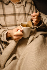 A cup of tea in the hands of a girl. The girl is drinking hot tea. A girl in pajamas is wrapped in a blanket and is drinking delicious tea. Enjoy the comforts of home. White cup close up in woman hand