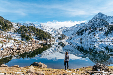 woman and reflection of the snowy mountains in the beautiful Baciver lake in the Pyrenees mountains of Val d'Aran (Aran Valley), Lleida, Catalonia, Spain - Powered by Adobe