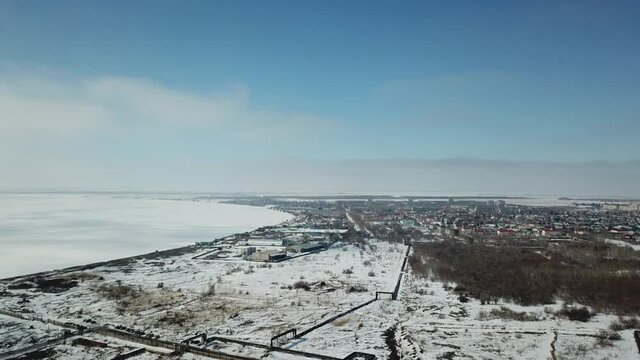 Forward moving aerial shot to snowed town when weather is sunny. Iced big water on the left of frame, some trees on the right. Blue sky with small clouds