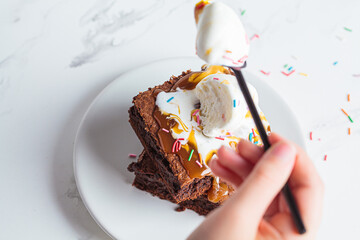 Children's hand eating brownie with scoop of ice cream, caramel and sprinkles, white background.