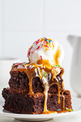 Stack of brownie squares with scoop of ice cream, caramel and sprinkles, white background.