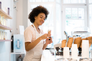 A beautiful mixed-race woman is smiling behind the stand of an ice cream parlor and serving the ice...